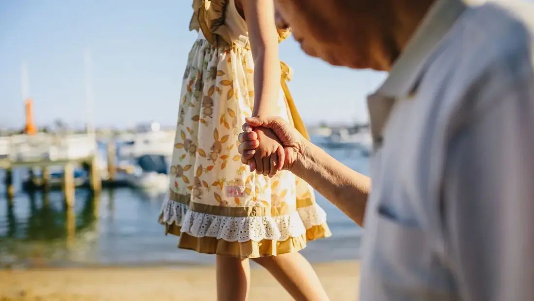 A child's hand held by an older person's hand, walking by the water